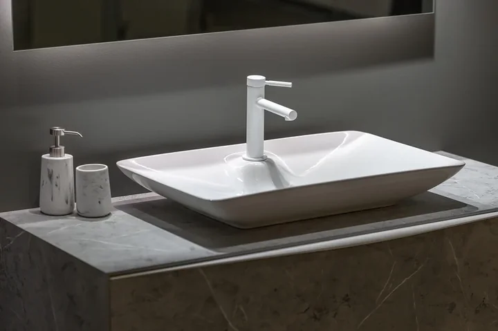 Biała prostokątna umywalka nablatowa z białą baterią, dozownikiem mydła i kubkiem na marmurowym blacie
