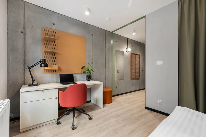Biurko z laptopem i czarną lampą, czerwone krzesło obrotowe, panel korkowy na betonowej ścianie, roślina doniczkowa.