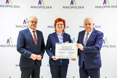 Trójka osób w garniturach stoi przed ścianką z logo Małopolska i wręcza symboliczny czek na modernizację stacji uzdatniania wody w Gromniku.
