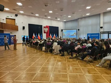 W auli odbywa się konferencja lub spotkanie z udziałem publiczności, na scenie stoją flagi Polski i UE, a na ekranie wyświetlana jest prezentacja.