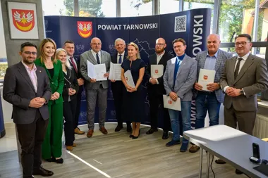 Grupa osób pozuje do zdjęcia podczas uroczystości w siedzibie powiatu tarnowskiego, kilku uczestników trzyma dokumenty z herbem powiatu.
