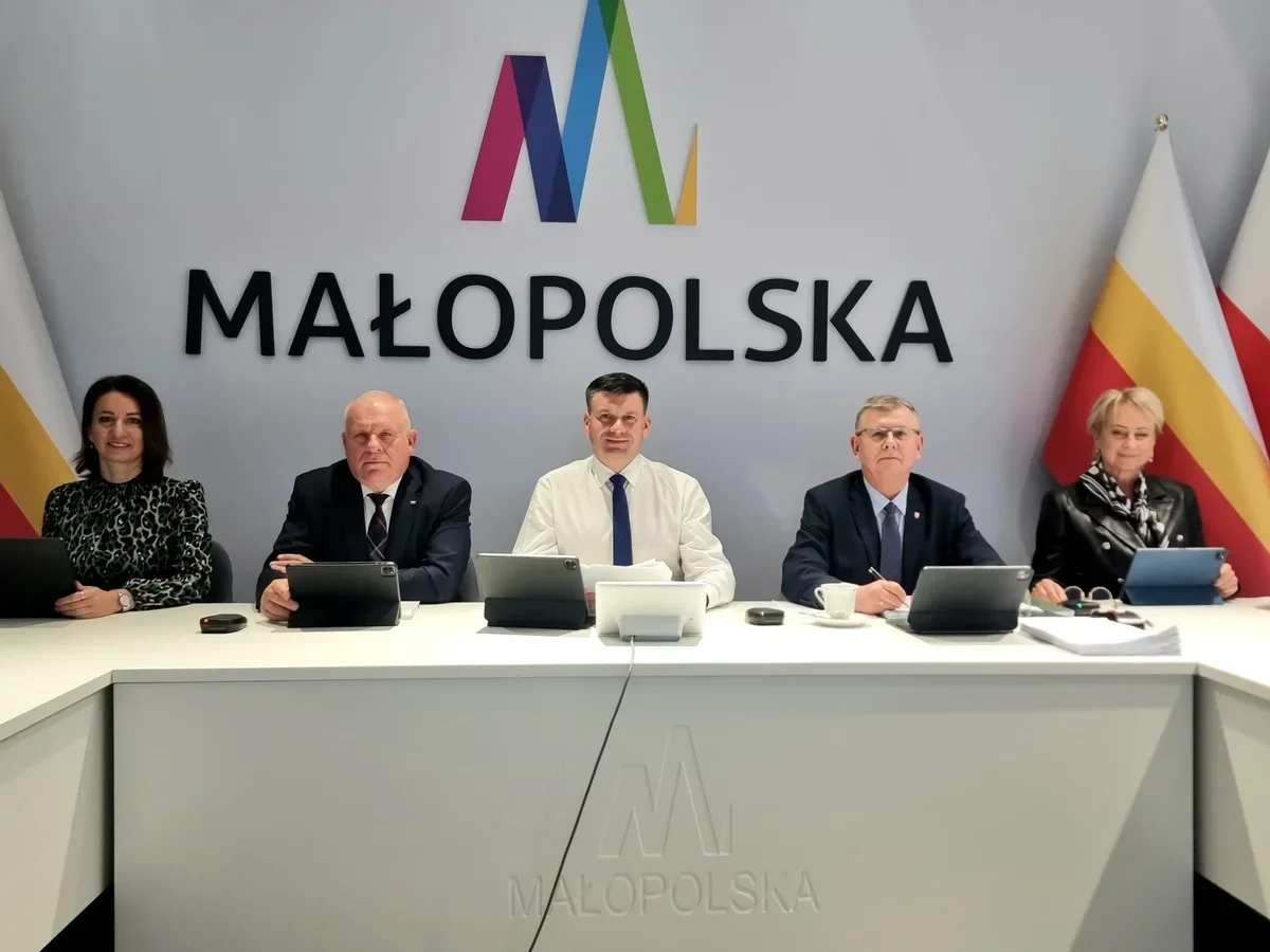 Pięć osób siedzi za białym stołem z tabletami podczas spotkania w sali konferencyjnej z logo Małopolska na ścianie.
